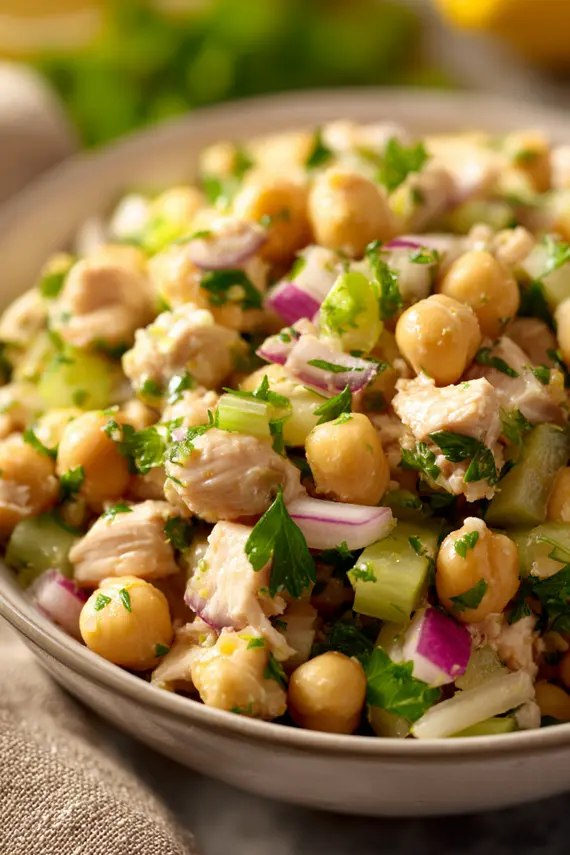 Freshly made chickpea chicken salad in a white serving bowl garnished with fresh herbs and lemon wedges on the side