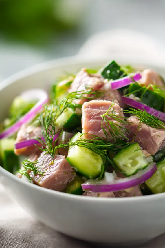 A beautifully plated tuna cucumber salad in a white bowl garnished with fresh dill and lemon wedges